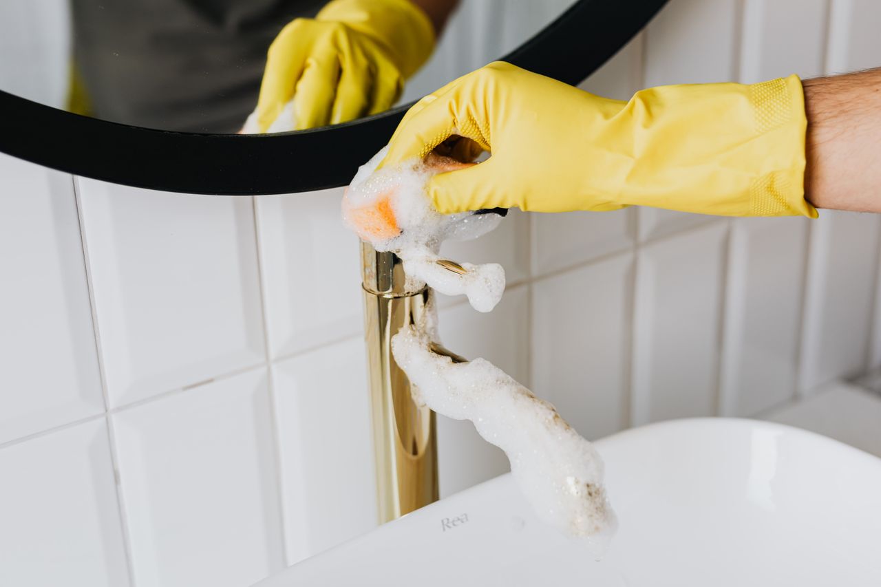 services-04 Personne anonyme portant des gants en latex jaunes et nettoyant un robinet doré avec une éponge dans une salle de bains moderne, pendant le ménage